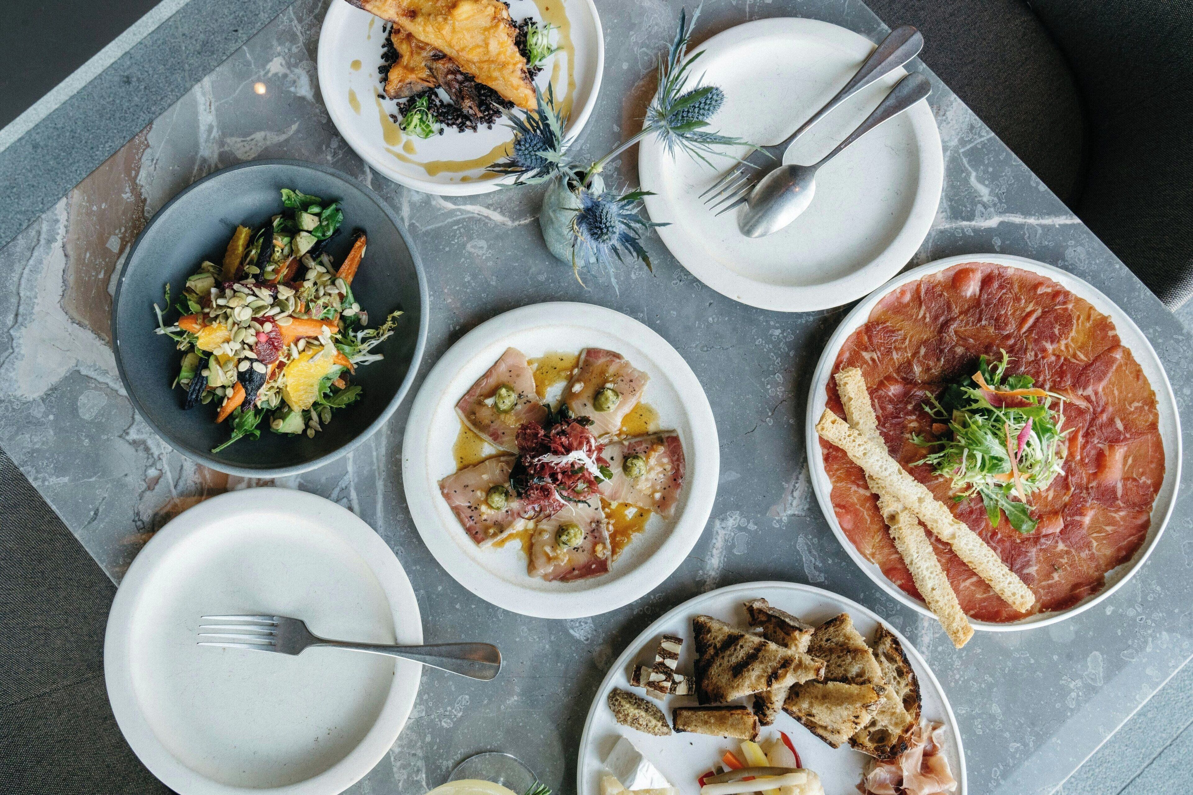 Une table à manger avec différents plats, notamment une assiette de jambon cru, une salade et du pain.