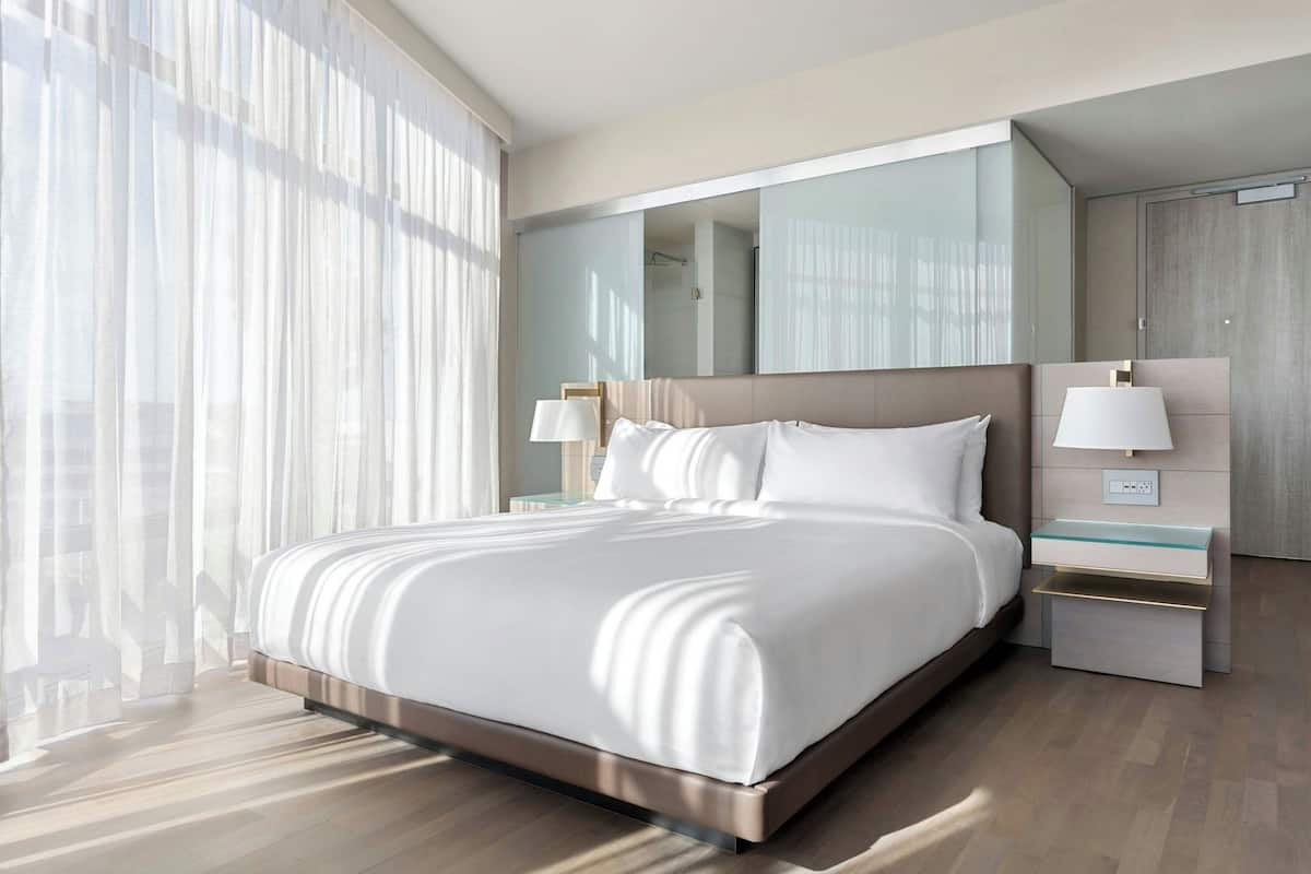 A modern hotel room with a large bed, a bedside table with a lamp, a mirror, and a wardrobe.