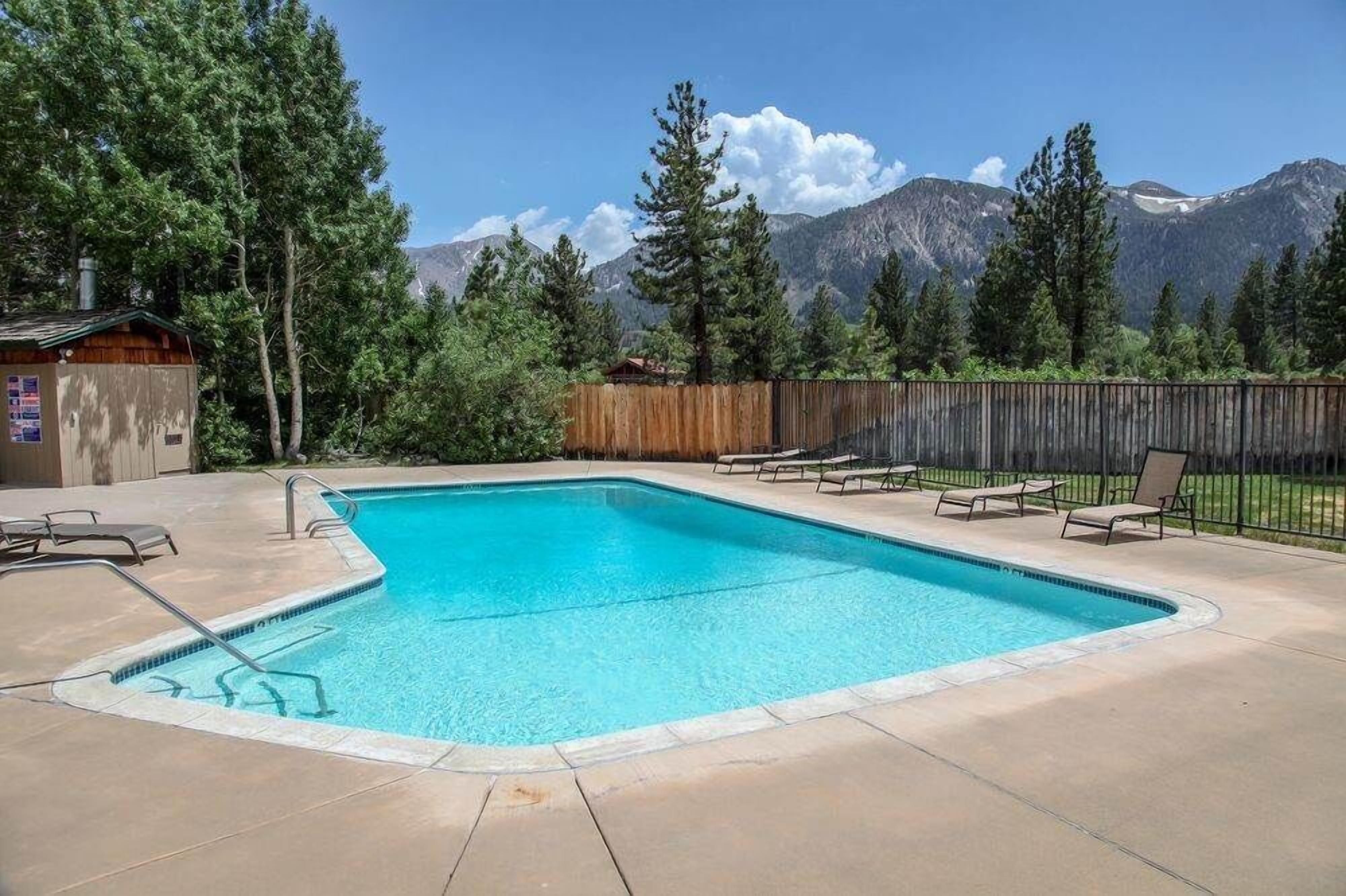 A rectangular swimming pool with a wooden deck, surrounded by a wooden fence and trees.