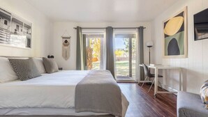 A bedroom with a large bed, a desk, and a chair. There is a window with curtains, a wall-mounted artwork, and a view of the outdoors.