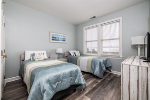 A bedroom with two beds, a bedside table with a lamp, a window with blinds, and a framed picture on the wall. - Sunset Island 53 Island Edge Drive (Ocean City)