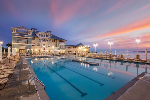 A swimming pool with lounge chairs and umbrellas, a building with multiple levels and balconies, and a sunset sky.