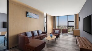 Une chambre d’hôtel moderne dotée d’une grande fenêtre, d’un canapé en cuir marron, d’une table basse ronde et d’un téléviseur à écran plat fixé au mur.