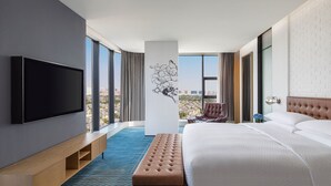 Een moderne hotelkamer met een groot bed, een flatscreen-tv, een bankje en uitzicht over de stad.