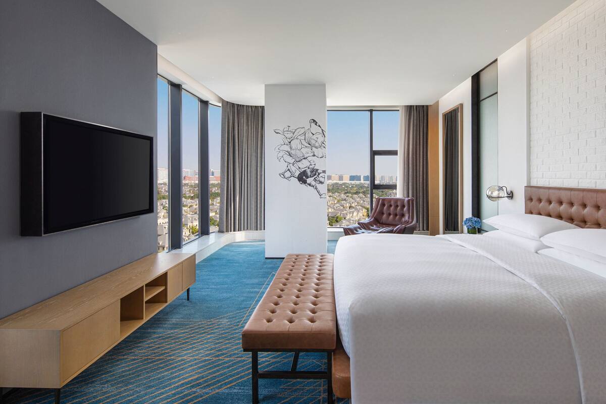 Ein modernes Hotelzimmer mit einem großen Bett, einem Flachbildfernseher, einer Sitzbank und Blick auf die Stadt.