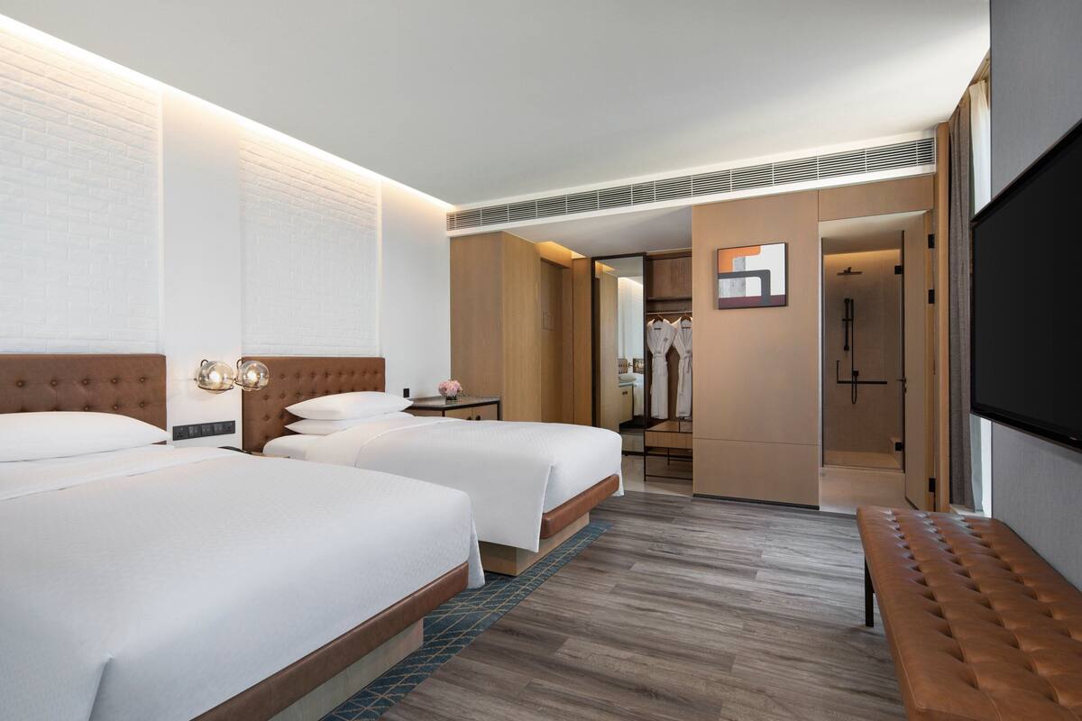 Ein Hotelzimmer mit zwei Betten, einer Bank, einem Fernseher, einem Spiegel und einem begehbaren Kleiderschrank.