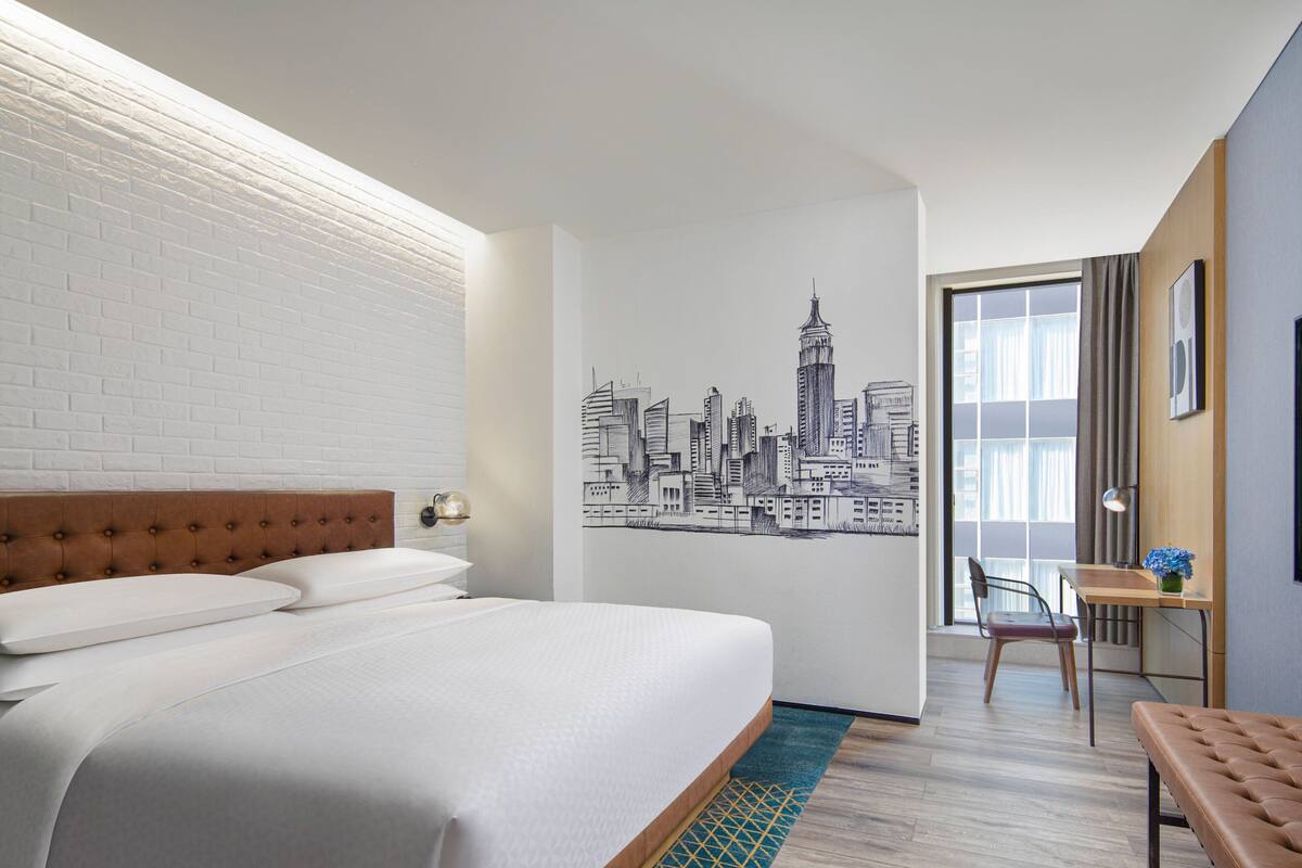 Ein Hotelzimmer mit einem großen Bett, einem Stadtbild-Wandgemälde, einem Schreibtisch mit Stuhl und einem fensterbepflanzten Fenster.