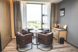 Una habitación de hotel moderna con una mesa redonda, cuatro sillas de cuero, una kitchenette pequeña y un amplio ventanal con cortinas.