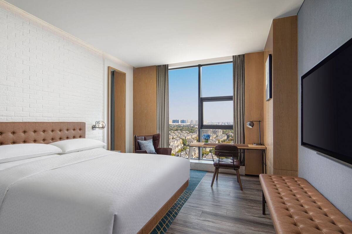 Ein Hotelzimmer mit einem großen Bett, einem Schreibtisch, einem Sessel, einer Bank und einem Fernseher.