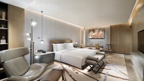 Una habitación de hotel moderna con una cama grande, una zona de estar con una silla y un banco, una mesita de noche, una lámpara de pie y una obra de arte grande en la pared.