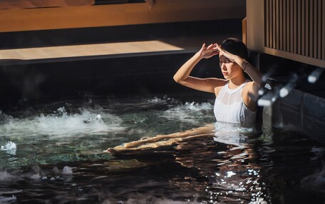 A person in a white tank top sitting in a hot tub with steam rising from the water.