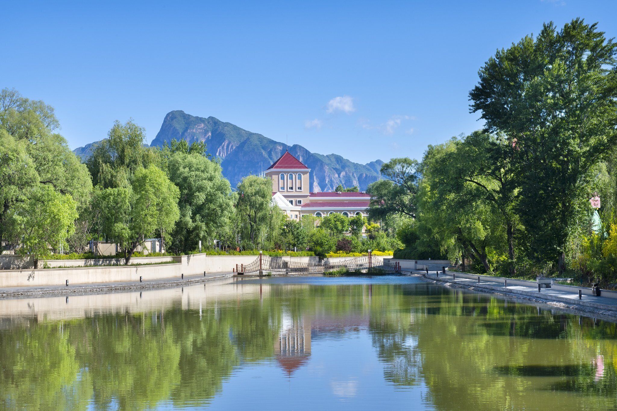 A serene lake with a reflection, surrounded by lush greenery and a prominent building with a red roof.