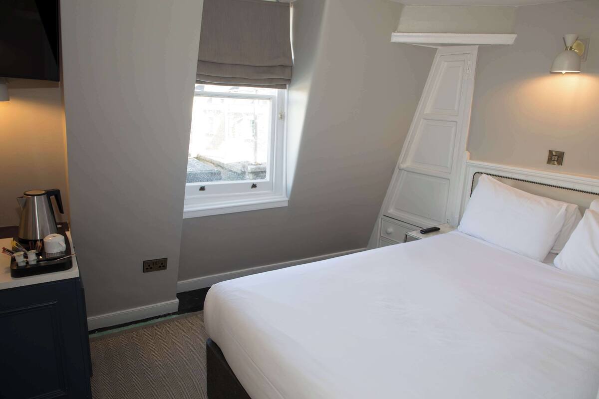 A hotel room with a bed, a desk with a coffee maker, a window with a view of the street, and a wall-mounted lamp.