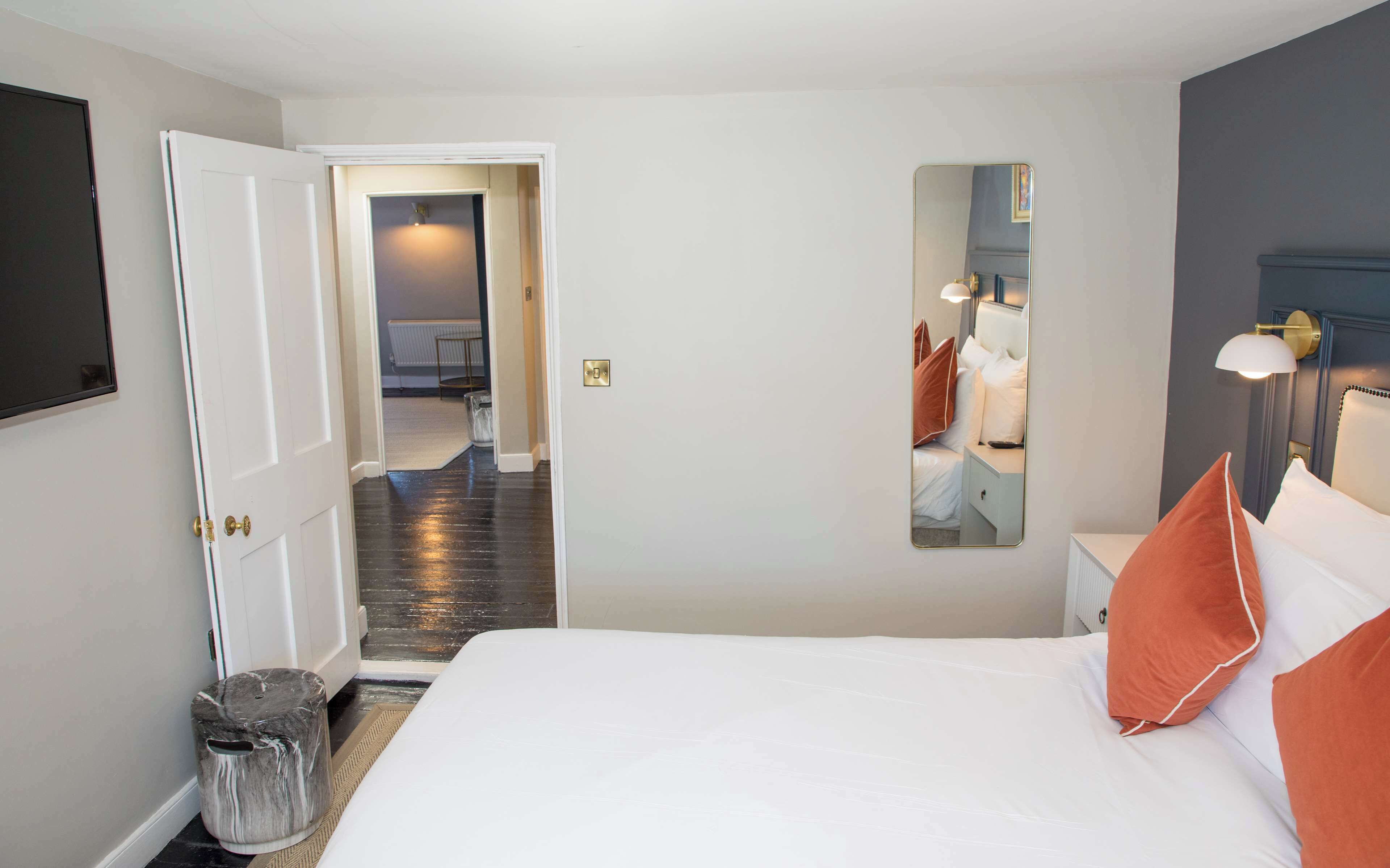 A hotel room with a bed, a mirror, a nightstand, a lamp, and a door leading to another room.