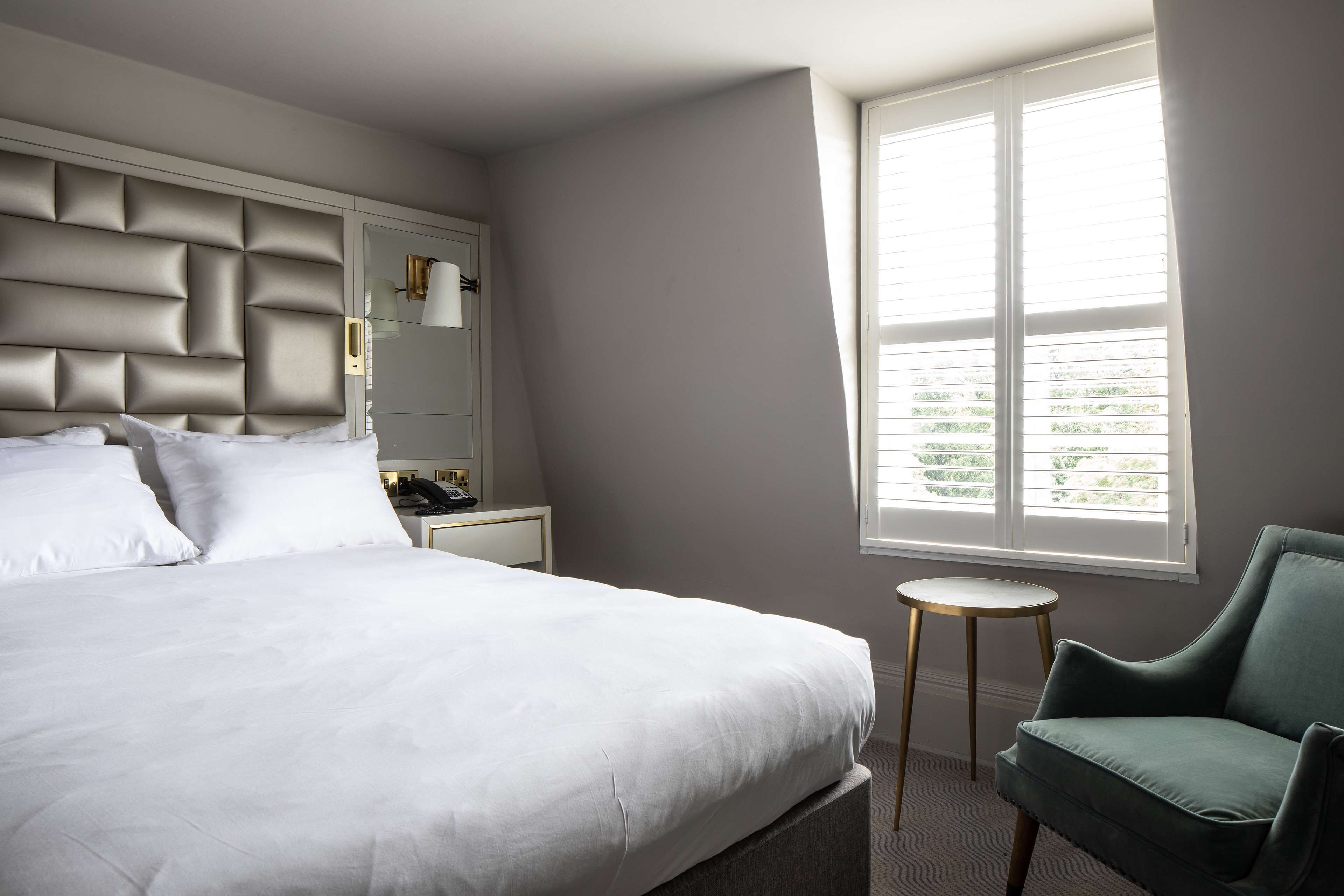 Una cama bien hecha con ropa de cama blanca, un cabecero tapizado en gris, una pequeña mesa redonda con base dorada y un sillón verde.