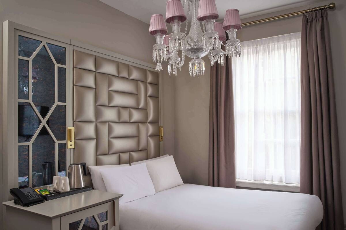 A hotel room with a large bed, a bedside table with a phone and coffee maker, a decorative wall panel, and a chandelier.