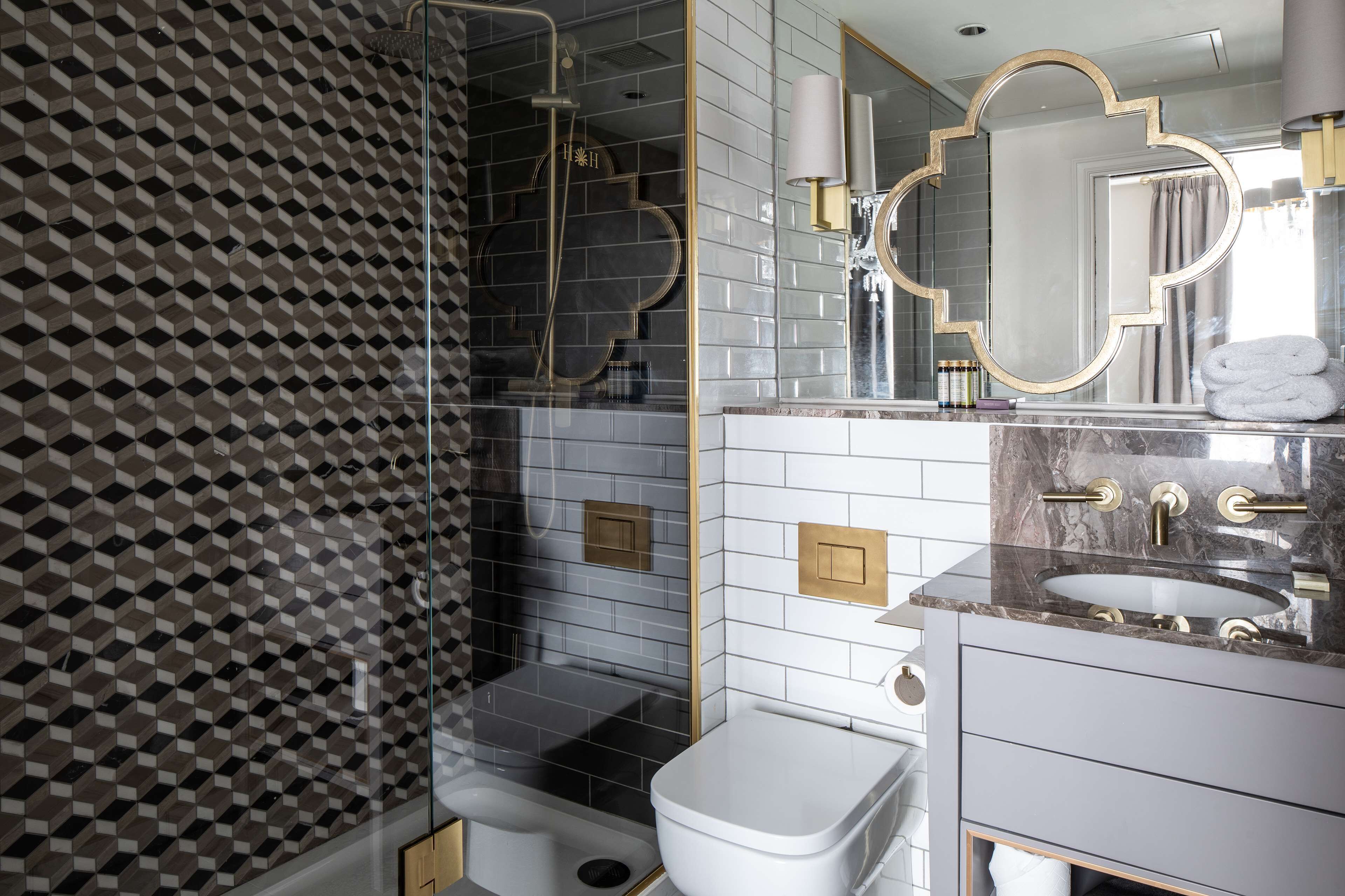 A modern bathroom with a geometric tile pattern, a glass-enclosed shower, a white toilet, a marble sink, and a vanity with gold fixtures.