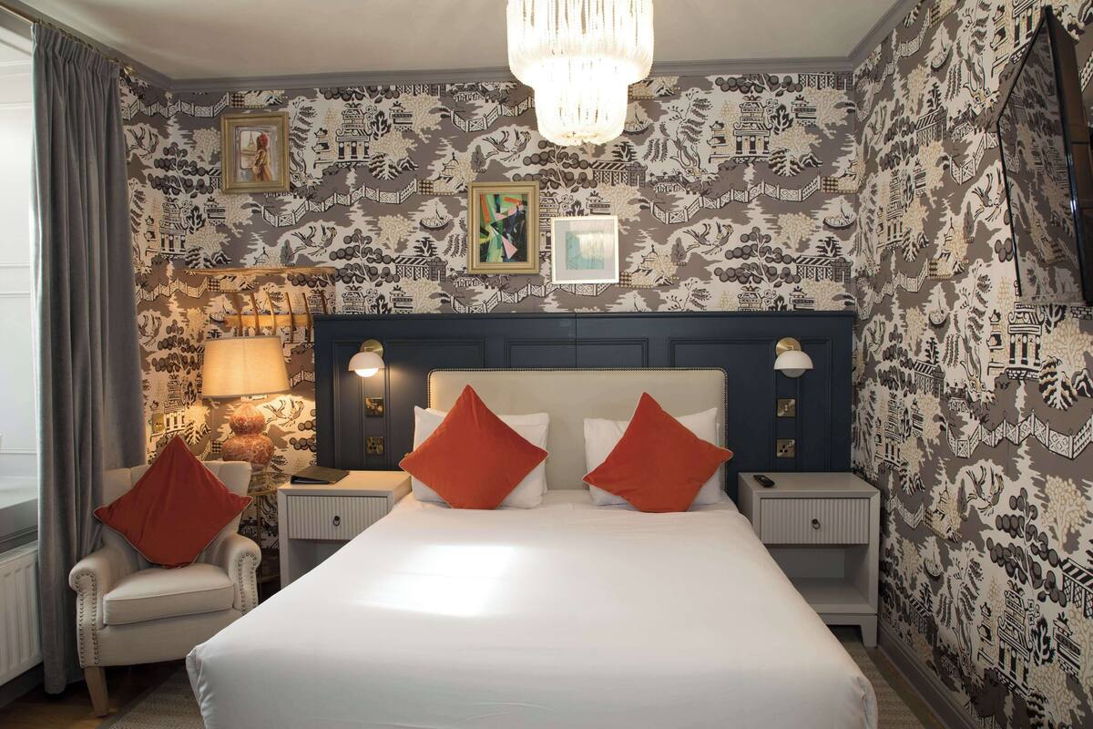A hotel room with a large bed, two nightstands, a chair, a lamp, and patterned wallpaper.