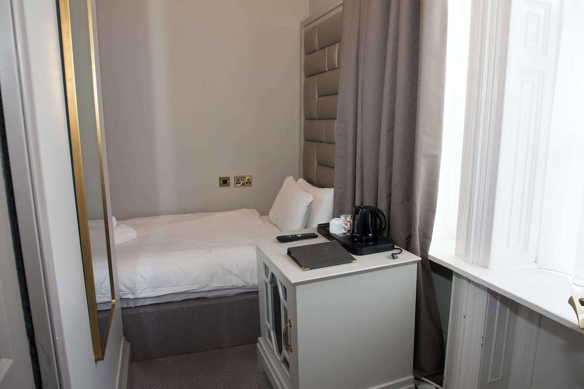 A small hotel room with a single bed, a bedside table with a remote control, a coffee maker, and a window with curtains.