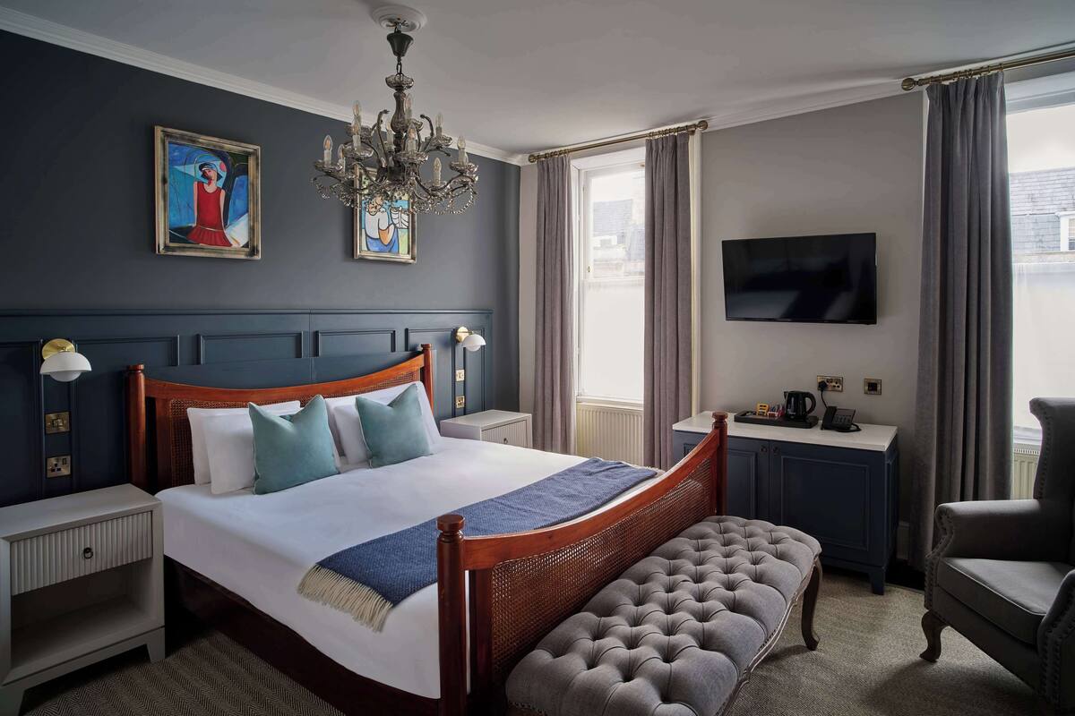 A bedroom with a large bed, a wooden headboard, a bench, a TV, a desk, and a chandelier.