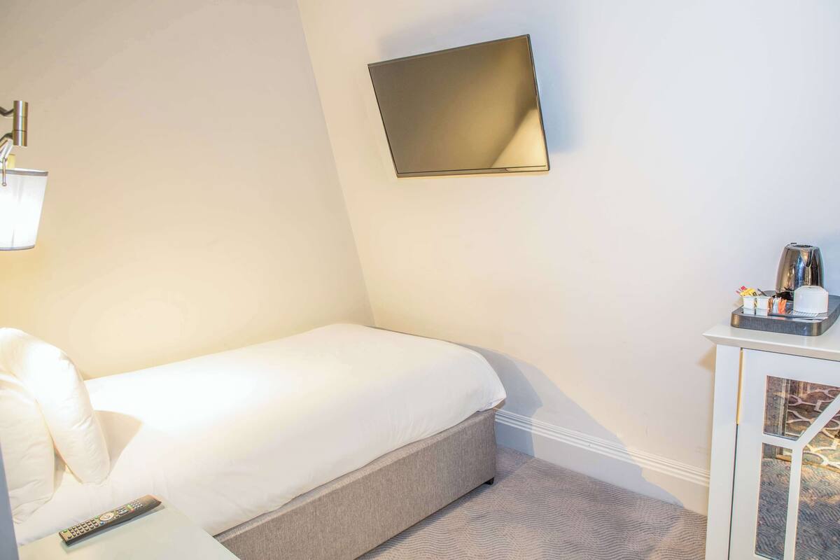 A hotel room with a bed, a bedside table with a remote control, a wall-mounted TV, and a small cabinet with toiletries.
