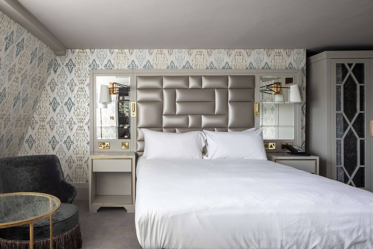A hotel room with a large bed, a dark armchair, a round table, and a nightstand with lamps.