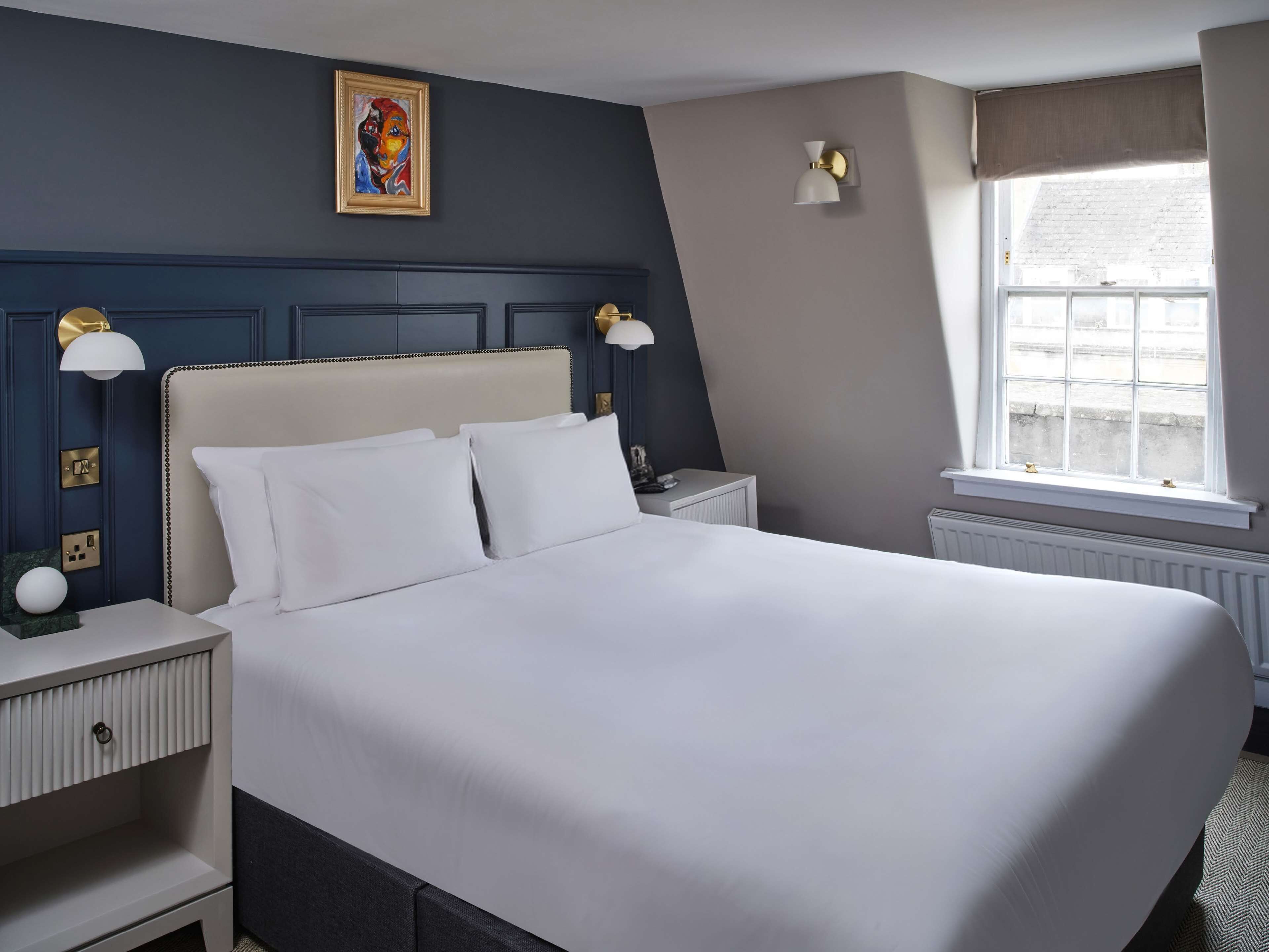 Siististi järjestetty hotellihuone, jossa on suuri sänky, yöpöydät, ikkuna verhoilla ja taulu seinällä.