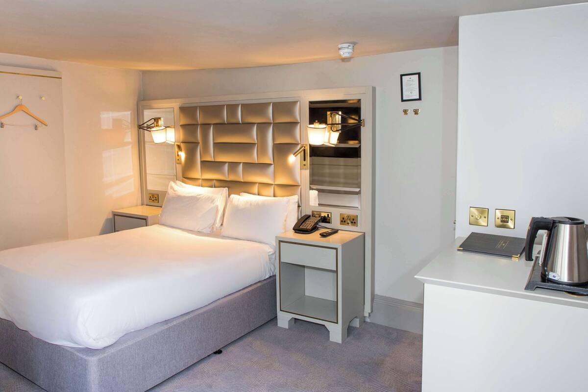 A hotel room with a large bed, bedside tables, a desk with a kettle, and a wardrobe.