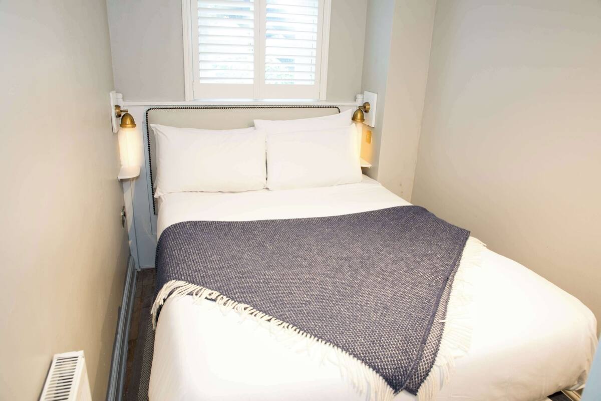 A neatly made bed with white linens and a dark blue blanket, a bedside table with a lamp, and a window with blinds.