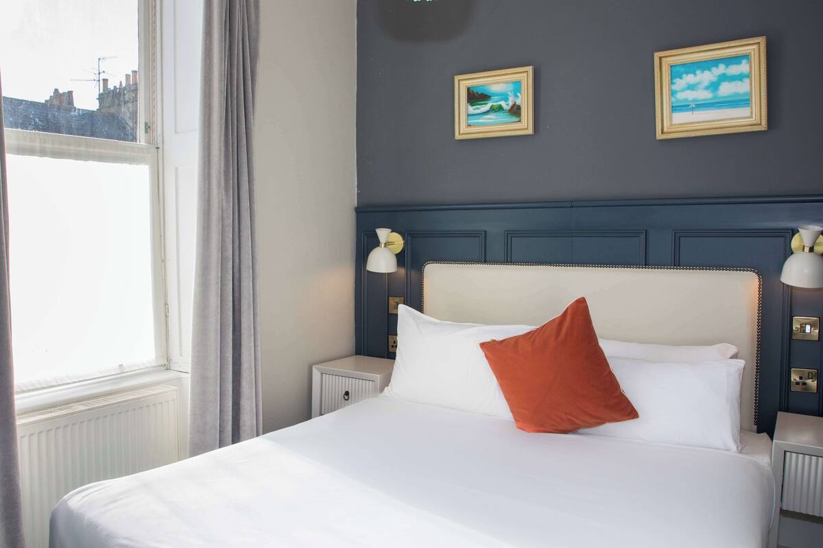A hotel room with a bed, two wall-mounted paintings, a window with curtains, and a nightstand with a lamp.