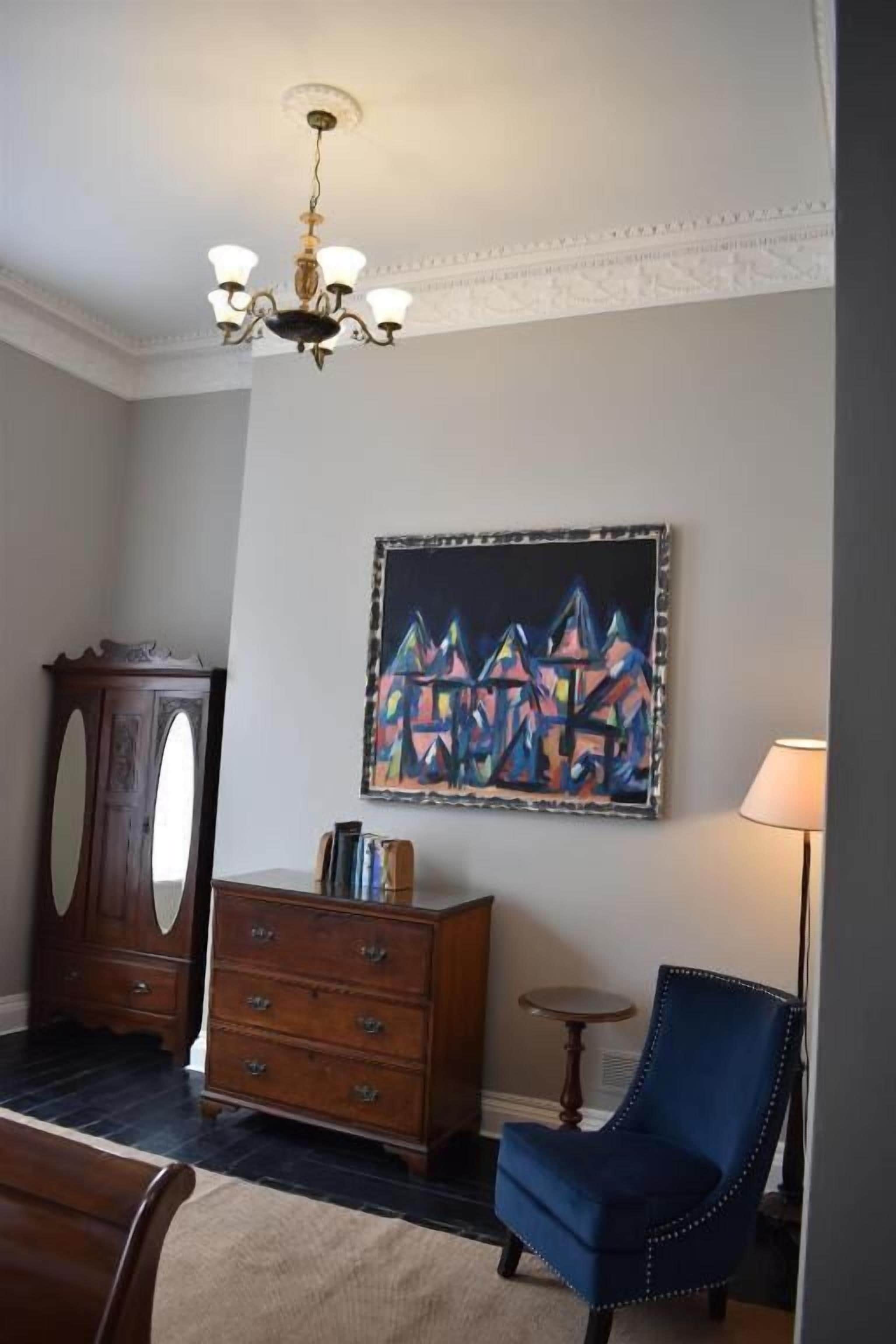 Una habitación con un aparador de madera, un sillón azul, un cuadro en la pared y una araña.