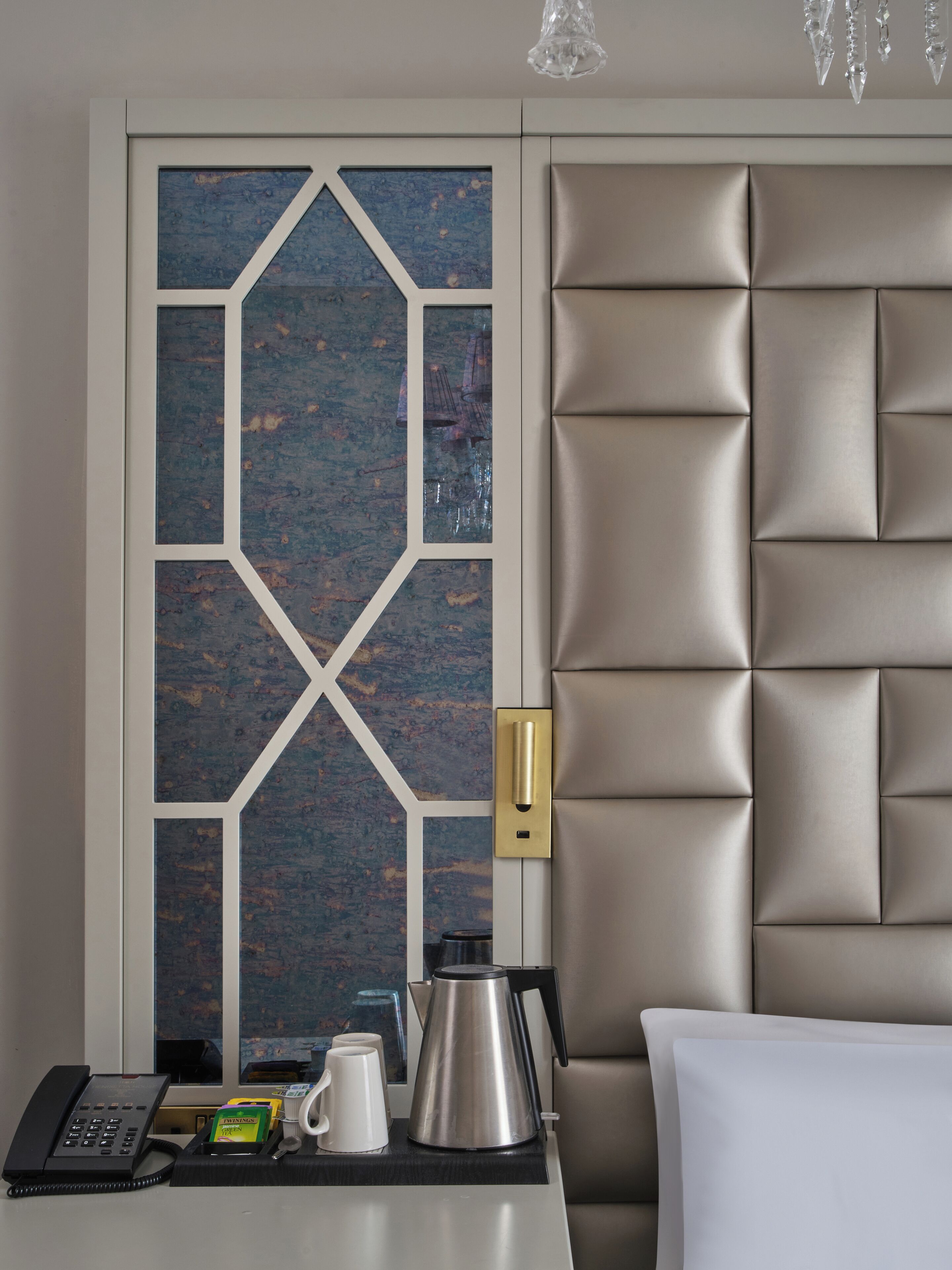 A hotel room with a modern design, featuring a geometric patterned wall, a tufted headboard, a bedside table with a phone, a kettle, and a coffee maker.