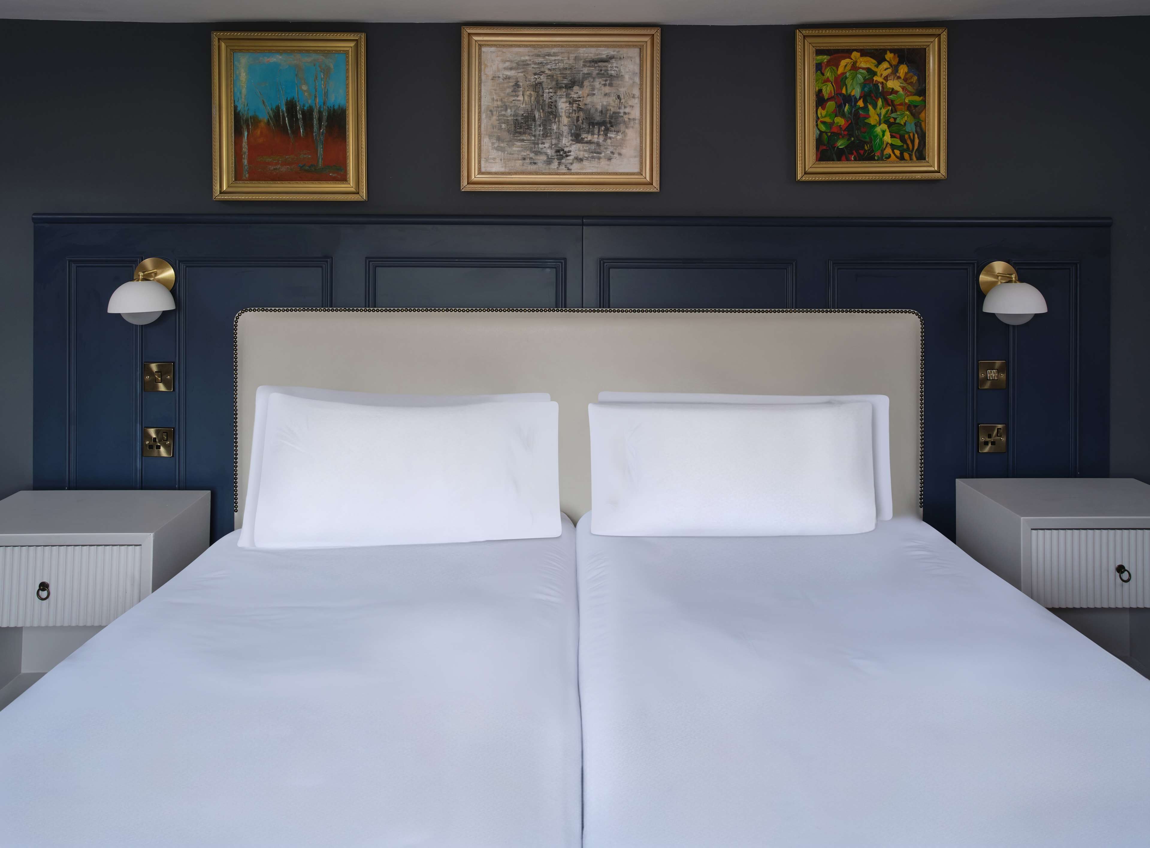 A double bed with white linens, a dark blue headboard, and three framed artworks above.