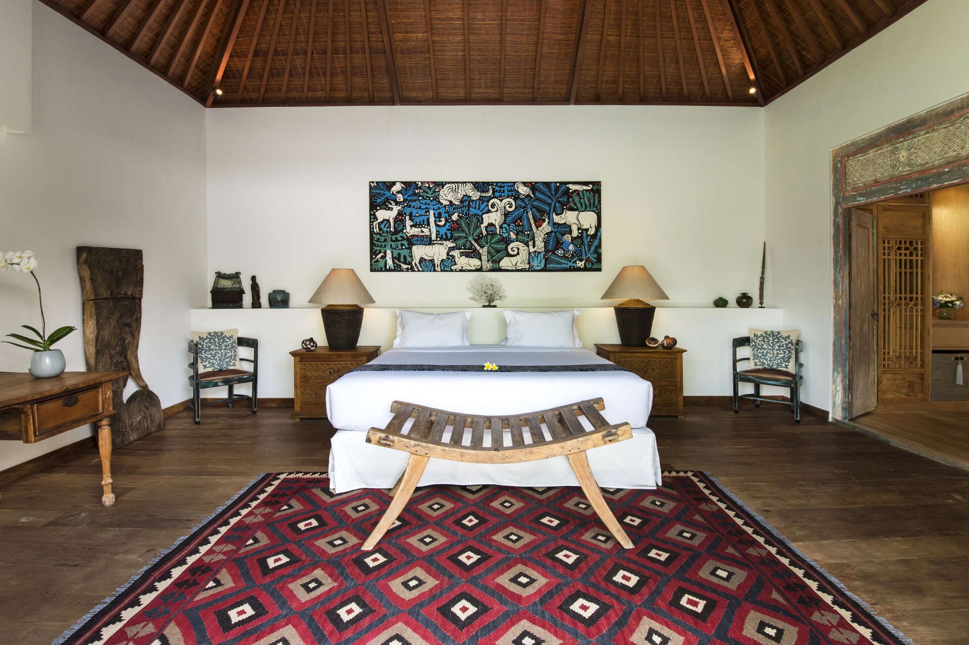 A bedroom with a large bed, two nightstands, a wooden bench, a patterned rug, and a wall painting.