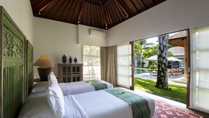 Une chambre d’hôtel avec deux lits, un plafond en bois, une fenêtre donnant sur une piscine et des palmiers, ainsi qu’une petite table avec une lampe.