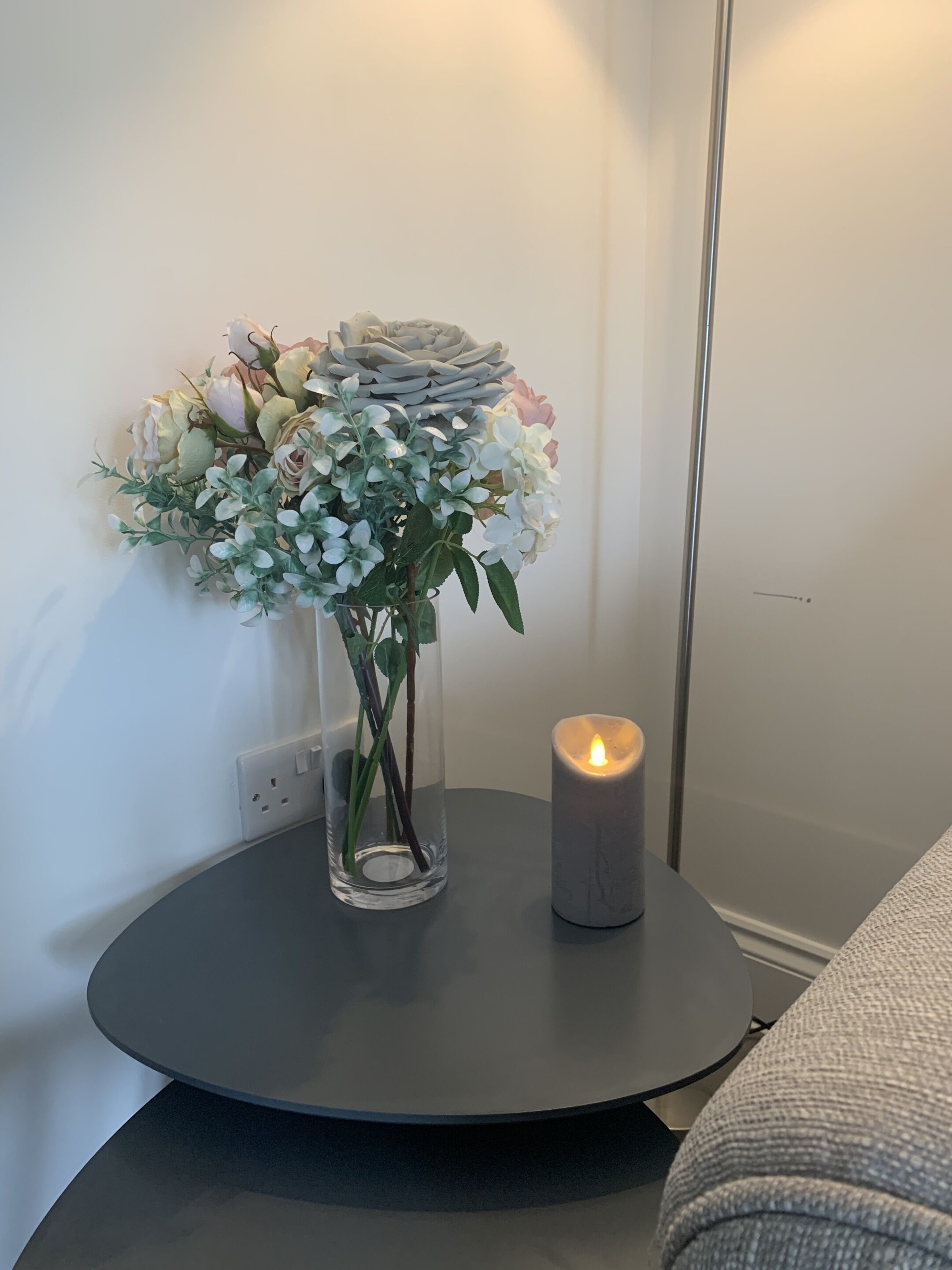 Een glazen vaas met kunstbloemen op een ronde tafel, naast een brandende kaars.