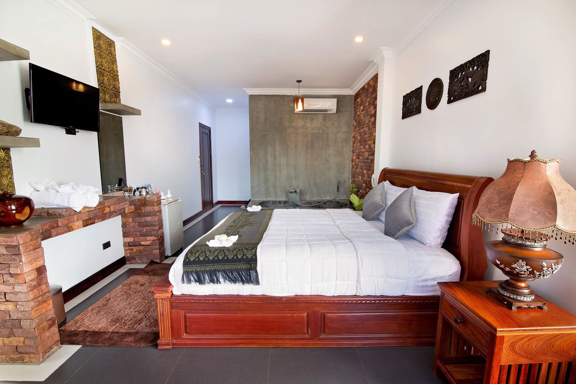 A hotel room with a large bed, a wooden nightstand, a TV mounted on the wall, and a brick accent wall.