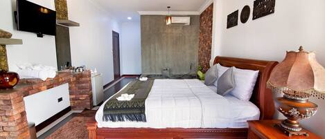A hotel room with a large bed, a wooden nightstand, a TV mounted on the wall, and a brick accent wall.