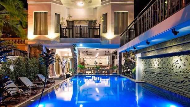 A swimming pool with a blue-lit interior, surrounded by lounge chairs and potted plants. - Damnak Villa Boutique (Siem Reap)