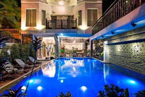 Ein Pool mit blau beleuchtetem Innenraum, umgeben von Liegestühlen und Topfpflanzen.