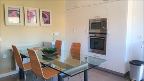 A modern kitchen with white cabinets, a microwave, and an oven. There is a glass dining table with wicker chairs and a bowl of fruit. - Villa André con Piscina, Barbacoa, Wifi & Satélite (Vícar)