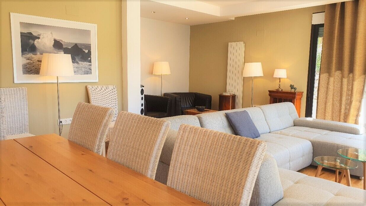 A modern living room with a wooden dining table, a grey sofa, and two armchairs. There are two floor lamps and a framed picture on the wall.