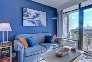 A living room with a grey sofa, blue cushions, a coffee table, and a large window.