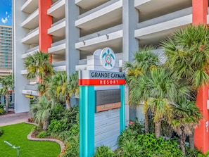 A hotel entrance with a sign for Grande Cayman Resort, a palm tree-lined driveway, and a modern building facade.