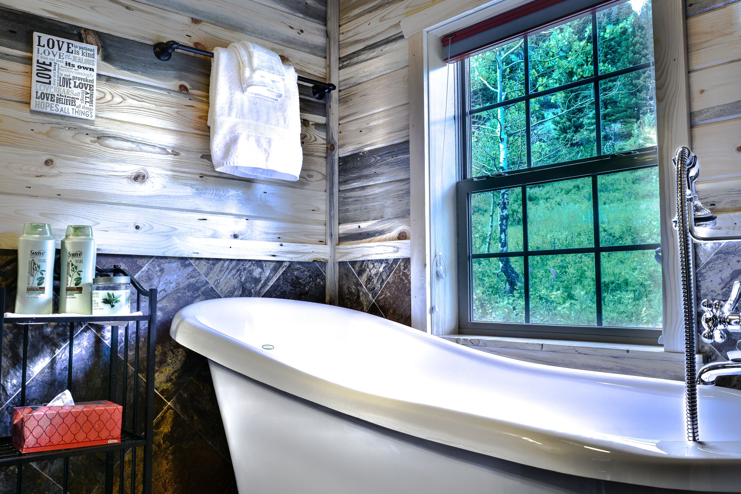A bathroom with a freestanding bathtub, a window with a view of greenery, a towel rack, and toiletries on a shelf.