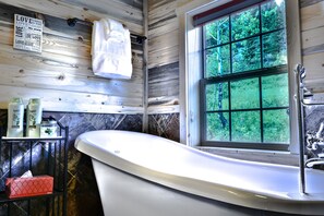 Un baño con una bañera independiente, una ventana con vistas a la vegetación, un perchero y artículos de aseo en un estante.