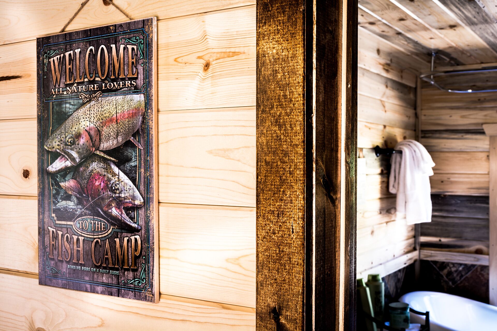 A wooden cabin interior with a welcome sign for a fish camp, a wooden wall, and a glimpse of a bathroom with a towel hanging.