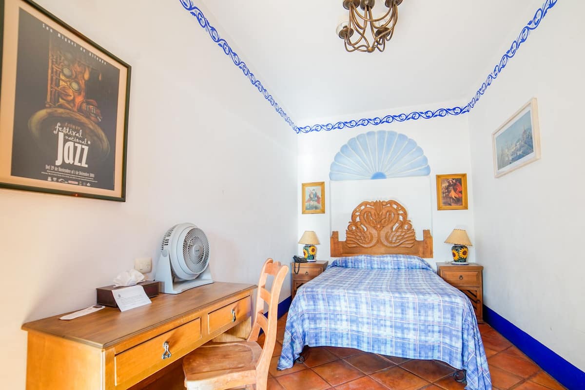 Ein Schlafzimmer mit einem Bett, einem Holztisch, einem Stuhl, zwei Nachttischen mit Lampen, einem Wandventilator und einem Poster an der Wand.