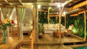 A room with a wooden floor, a bed, a sofa, a wicker chair, a small table, and a decorative vase with flowers.