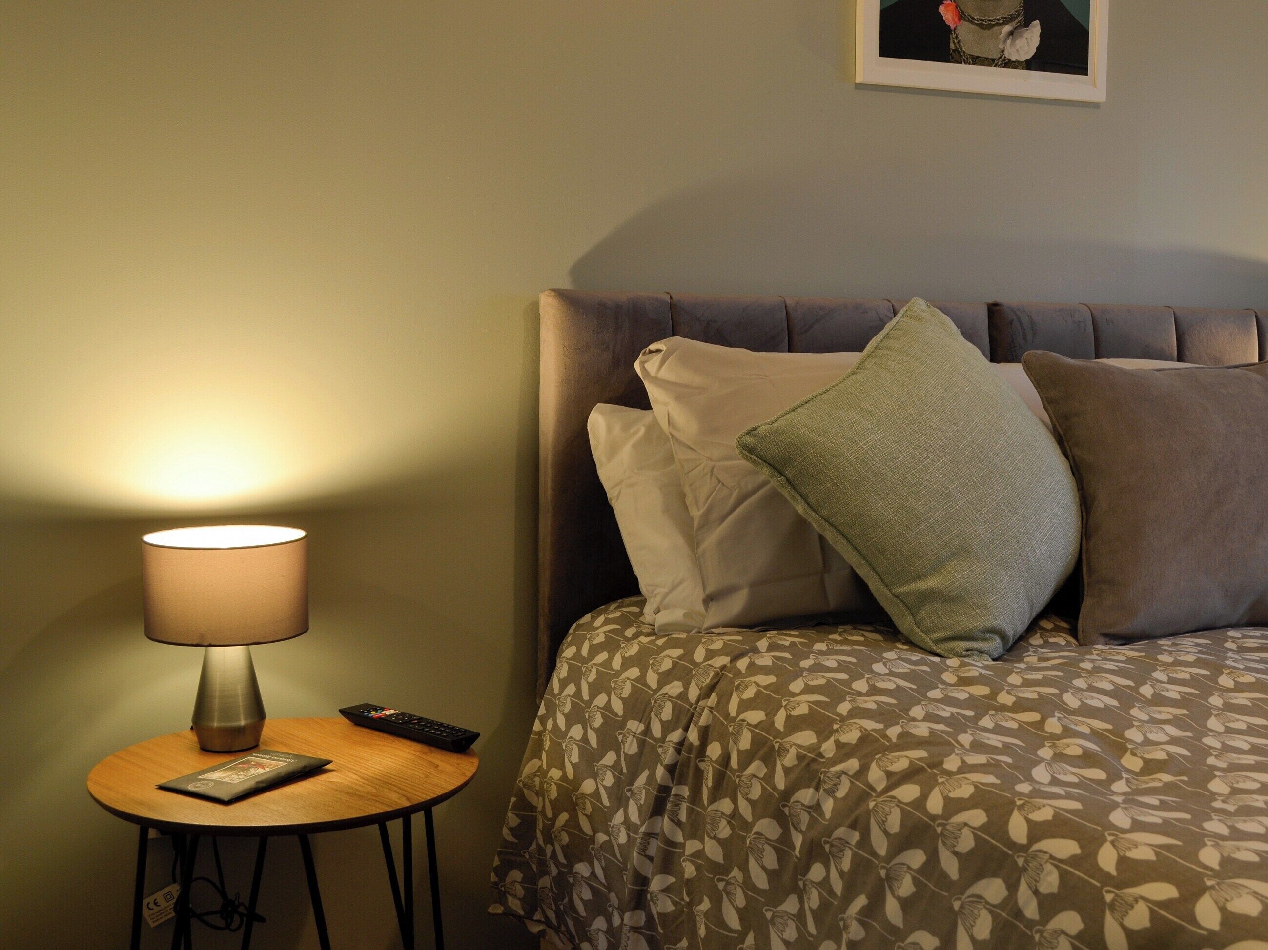 A neatly made bed with patterned bedding, a bedside lamp, a remote control, and a small table with a book.